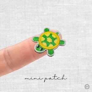 Könnte beinhalten: Ein kleiner, gestickter Schildkrötenaufnäher mit gelb-grünem Panzer, grünen Gliedmaßen und einem weißen Rand. Der Text "mini patch" ist in schwarzer Schrift darunter geschrieben. Der Aufnäher wird auf einem Finger gezeigt.