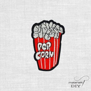 Könnte beinhalten: Gestickter Aufnäher mit einem Popcorn-Design. Der rote Popcorn-Behälter hat weiße vertikale Streifen und den weißen Schriftzug "POP CORN". Das Popcorn selbst ist weiß, und der Aufnäher hat einen schwarzen Rand.