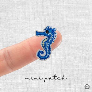 Könnte beinhalten: Ein blauer Mini-Patch in Form eines Seepferdchens mit gelben Akzenten und schwarzen Augen. Der gestickte Patch hat einen weißen Rand und den Schriftzug "mini patch" in schwarzer Schrift.