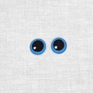 Könnte beinhalten: Gesticktes Paar Cartoon-Augen mit schwarzen Pupillen, weißen Glanzlichtern und blauen Umrissen. Die Augen sind vor einem weißen, strukturierten Stoffhintergrund platziert. Diese Aufnäher könnten für Bastelarbeiten oder zum Verschönern von Kleidung verwendet werden.