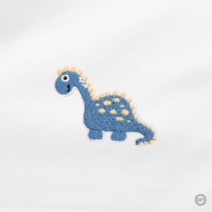 Könnte beinhalten: Ein gestickter Aufnäher, der einen blauen Dinosaurier mit gelben Flecken entlang des Rückens und des Schwanzes zeigt. Der Dinosaurier hat ein weißes Auge mit schwarzer Pupille und ein kleines Lächeln. Der Aufnäher liegt auf weißem Grund mit DIY-Logo.