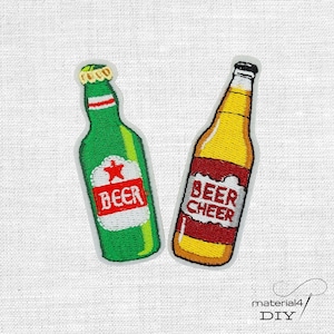Könnte beinhalten: Zwei gestickte Aufbügler in Form von Bierflaschen. Der linke Aufnäher ist grün mit einem weißen Etikett, einem roten Stern und dem Schriftzug BEER. Der rechte Aufnäher ist gelb mit einem roten Etikett und dem Schriftzug BEER CHEER in Weiß.