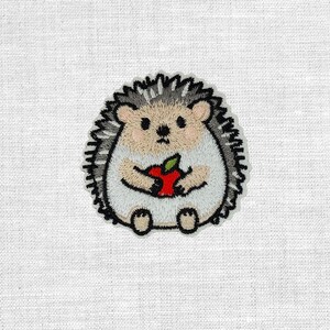Könnte beinhalten: Gestickter Aufnäher mit einem niedlichen Igel, der einen roten Apfel hält. Der Igel hat ein hellbraunes Gesicht und einen hellbraunen Bauch mit grauen und schwarzen Stacheln. Der Aufnäher ist schwarz umrandet und befindet sich auf weißem Hintergrund.