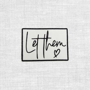 Könnte beinhalten: Ein rechteckiger Aufnäher mit schwarzem Rand und den Worten "Let them" in handschriftlichem Stil, mit einem kleinen Herzsymbol. Der Aufnäher befindet sich auf einem weißen Stoffhintergrund.
