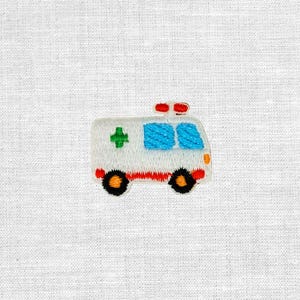 Könnte beinhalten: Ein gestickter Aufnäher in Form eines Krankenwagens. Das Fahrzeug ist weiß mit einem grünen Kreuz an der Seite, blauen Fenstern, einem roten Streifen am unteren Rand und zwei schwarzen Rädern. Auf dem Dach sind rote Lichter und seitlich ein orangefarbenes Licht.