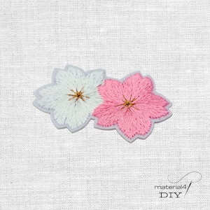 Könnte beinhalten: Gestickter Blumenaufnäher mit zwei Blüten, eine weiße und eine rosa, mit gelben Zentren. Die Aufnäher sind hellgrau umrandet und vor einem weißen Stoffhintergrund platziert. Der Text "material4 DIY" befindet sich in der unteren rechten Ecke.