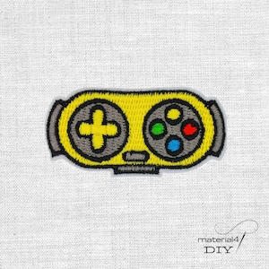 Könnte beinhalten: Gestickter gelber Videospiel-Controller-Patch mit schwarzen und grauen Details. Der Controller hat ein Steuerkreuz, farbige Knöpfe und eine schwarze Umrandung. Der Patch befindet sich auf einem weißen Stoffhintergrund.