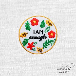 Könnte beinhalten: Ein kreisförmiger gestickter Aufnäher auf weißem Stoffhintergrund. Der Aufnäher hat eine weiße Mitte mit dem schwarzen Text I AM enough, umgeben von roten Blumen, grünen Blättern und gelben herbstfarbenen Blättern, eingefasst mit Goldfaden.