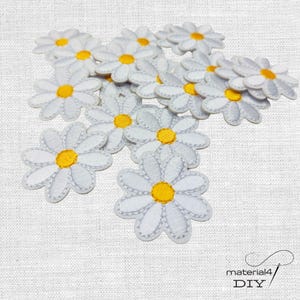 Könnte beinhalten: Eine Ansammlung kleiner, weißer Gänseblümchen-Aufnäher mit leuchtend gelben Mitten. Jeder Aufnäher verfügt über gestickte Blütenblätter und einen kreisförmigen gelben Kern, die zur Applikation auf Stoff gedacht sind. Der Text material4 DIY ist sichtbar.
