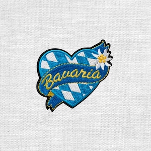 Könnte beinhalten: Gestickter Aufnäher in Form eines blauen Herzens mit einem weiß-blauen Rautenmuster. Ein blaues Banner mit dem Wort "Bavaria" in goldener Schrift verläuft über das Herz. Auf der rechten Seite des Herzens befindet sich eine weiße Blume mit gelbem Zentrum.
