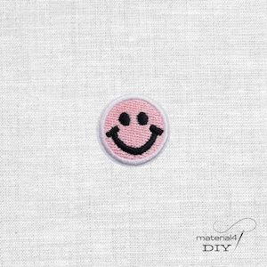 Könnte beinhalten: Ein kreisförmiger gestickter Aufnäher mit einem hellrosa Smiley-Gesicht, zwei schwarzen runden Augen und einem schwarzen geschwungenen Mund. Der Aufnäher hat einen weißen gestickten Rand und ist auf einem weißen Stoff mit Textur zentriert, inklusive Text material4 DIY.