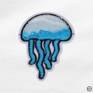Könnte beinhalten: Ein Quallen-Aufnäher mit Pailletten in Weiß, Hellblau und Blau. Die Qualle hat eine abgerundete Oberseite mit hellblauen und weißen Pailletten sowie blaue Pailletten-Tentakel mit schwarzen Stickkonturen.