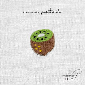 Könnte beinhalten: Ein gestickter Mini-Aufnäher in Form einer Kiwischeibe mit brauner, flauschig strukturierter Außenseite, hellgrünem Inneren, schwarzen Kernen und weißer Mitte. Der Text "mini patch" steht darüber, "material4 DIY" steht darunter.