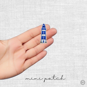 Könnte beinhalten: Ein kleiner gestickter Aufnäher mit einem Leuchtturm-Motiv in blauen und weißen Querstreifen, einem dunkelblauen oberen Bereich und einem weißen Sockel. Der Leuchtturm ist mit einer einfachen architektonischen Struktur und einem kleinen Torbogen dargestellt.
