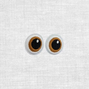 Könnte beinhalten: Gestickte Augen-Patches mit weißem Hintergrund, braunen Iris und schwarzen Pupillen. Diese Patches sind zum Aufnähen auf Stoff gedacht. Sie eignen sich ideal, um Kleidung, Accessoires oder Bastelprojekten eine skurrile Note zu verleihen.