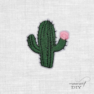 Könnte beinhalten: Gestickter grüner Kaktus-Aufnäher mit schwarzen Stacheln und einer rosa Blüte. Der Kaktus hat ein detailliertes Stichdesign und ist vor einem weißen Hintergrund platziert. Der Aufnäher ist ein DIY-Bastelartikel.