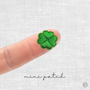 Könnte beinhalten: Ein kleiner, gestickter grüner vierblättriger Kleeblatt-Aufnäher. Der Aufnäher wird auf einem Finger gehalten, darunter steht in schwarzer Schrift "mini patch". Der Aufnäher ist ein DIY-Bastelartikel.