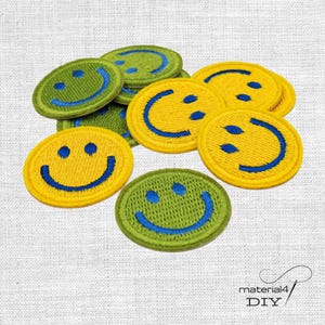 Könnte beinhalten: Eine Sammlung runder, bestickter Aufnäher mit Smiley-Gesichtern. Die Aufnäher sind in leuchtendem Gelb und Grün gehalten und zeigen jeweils ein blau gesticktes Smiley-Gesicht mit zwei runden Augen und einem geschwungenen Mund auf strukturiertem Stoff.