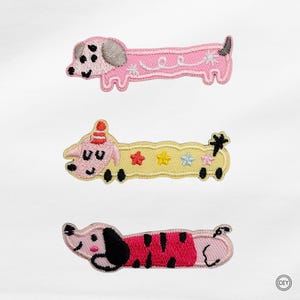 Könnte beinhalten: Drei gestickte Aufnäher in Form von langen Hunden. Der obere Aufnäher ist rosa mit einem grauen Ohr und weißen Wirbeldetails. Der mittlere Aufnäher ist gelb mit einem Partyhut und bunten Sternen. Der untere Aufnäher ist rosa mit schwarzen Flecken.