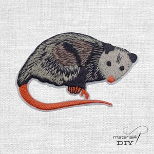 Könnte beinhalten: Gestickter Opossum-Aufnäher mit detaillierten grauen, schwarzen und braunen Fellzeichnungen, einer rosa Nase und einem orangefarbenen Schwanz. Der Aufnäher befindet sich auf einem weißen Stoffhintergrund. Der Text "material4 DIY" befindet sich unten rechts.