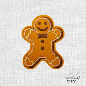Könnte beinhalten: Ein Lebkuchenmann-Applikation in Braun- und Beigetönen, mit einem lächelnden Gesicht, einer Fliege und Knopfformen. Die Applikation ist wahrscheinlich für DIY-Projekte wie Nähen oder Basteln gedacht.