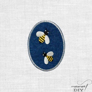 Könnte beinhalten: Ein ovaler Aufnäher aus Jeansstoff mit einem gestickten Design von zwei Bienen mit gelb-schwarz gestreiften Körpern und weißen Flügeln. Der Aufnäher ist mit einem weißen gestickten Rand um den gesamten Umfang der ovalen Form versehen.