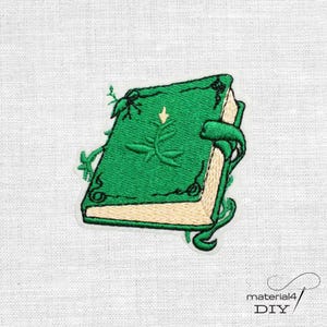 Könnte beinhalten: Ein gestickter Aufnäher, der ein grünes Buch mit hellgelbem Seitenrand zeigt. Der Buchdeckel ist mit einem dunkelgrünen Pflanzenmotiv verziert und teilweise von grünen Ranken umgeben. Das Design enthält ein kleines Insekt.