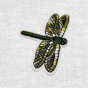 Könnte beinhalten: Gestickter Libellen-Aufnäher mit weißem Rand. Die Libelle hat gelbe und grüne Flügel mit detaillierten Nähten, einen dunkelgrünen Körper und einen weißen Hintergrund.