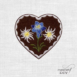 Könnte beinhalten: Ein herzförmiger Aufnäher aus dunkelbraunem Stoff mit weißem Wellenrand. In der Mitte befindet sich eine gestickte blaue Blume mit gelber Mitte, flankiert von zwei weißen Edelweißblüten mit gelben Zentren und grünen Stielen.
