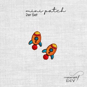Könnte beinhalten: Zwei gestickte Raketen-Aufnäher mit orangefarbenen Körpern, gelben Spitzen, blauen Flügeln und roten Auspuffflammen. Der Text mini patch 2er Set steht über den Raketen und der Text material4 DIY befindet sich in der unteren rechten Ecke.
