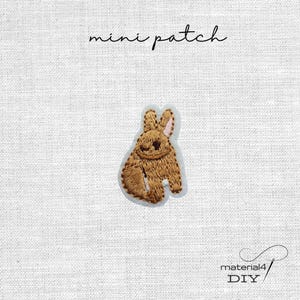 Könnte beinhalten: Ein brauner, gestickter Hasenaufnäher mit rosa Innenohren und einem dunkelbraunen Auge. Der Text "mini patch" steht über dem Hasen und der Text "material4 DIY" befindet sich in der unteren rechten Ecke auf einem weißen Stoffhintergrund.