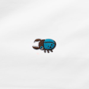 Könnte beinhalten: Gestickter Aufnäher mit einem stilisierten Käfer-Design. Der Käfer hat einen blauen Körper mit kleinen schwarzen Punkten und einen braunen Kopf und Zangen. Geeignet für Kleidung oder Accessoires.