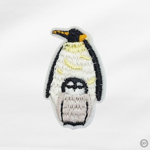 Könnte beinhalten: Gestickter Aufnäher mit einem Pinguin und seinem Küken. Der Pinguin hat einen schwarzen Kopf, einen gelb-schwarzen Schnabel und einen weißen und gelben Körper. Das Küken ist grau und schwarz. Der Aufnäher befindet sich auf weißem Hintergrund.