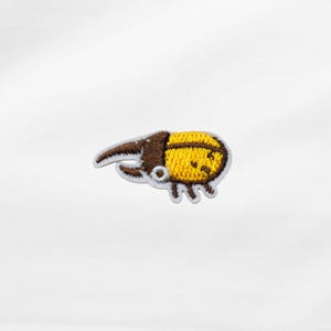 Könnte beinhalten: Gestickter Aufnäher mit einem Cartoon-Käfer-Design. Der Käfer hat einen gelben Körper mit braunen Akzenten und einem lächelnden Gesicht. Der Aufnäher ist für Kleidung oder Accessoires gedacht.