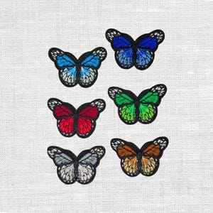 Könnte beinhalten: Sechs gestickte Schmetterlingsaufnäher in verschiedenen Farben: Blau, Rot, Grün, Grau und Braun. Jeder Schmetterling hat eine schwarze Umrandung und detaillierte Flügelmuster. Die Aufnäher befinden sich auf einem weißen Stoffhintergrund.