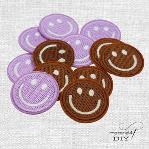 Könnte beinhalten: Eine Sammlung runder gestickter Aufnäher mit Smiley-Motiven in Helllila und Braun. Jeder Aufnäher zeigt einen gestickten weißen lächelnden Mund und zwei weiße kreisförmige Augen auf einem einfarbigen Hintergrund.