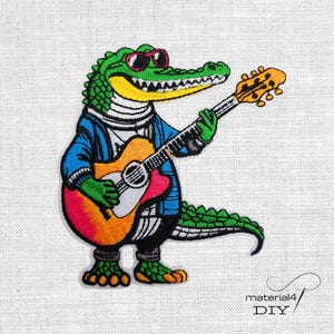 Könnte beinhalten: Gestickter Aufnäher mit einem grünen Alligator, der eine Sonnenbrille und eine blaue Jacke trägt und eine bunte Akustikgitarre spielt. Der Alligator hat einen gelben Bauch und steht auf zwei Beinen. Der Aufnäher befindet sich auf weißem Hintergrund.