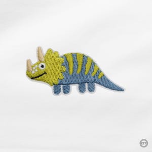 Könnte beinhalten: Ein gestickter Aufbügler in Form eines Triceratops-Dinosauriers. Der Dinosaurier hat einen hellgelben Kopf mit zwei Hörnern und einen blauen Körper, der mit vertikalen gelben Streifen verziert ist. Der Aufnäher hat einen weißen Rand und ein kleines DIY-Logo.