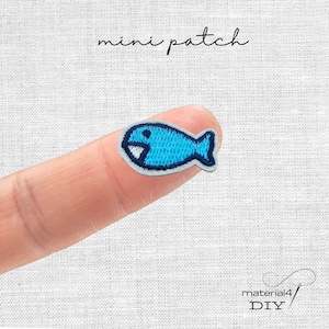 Könnte beinhalten: Ein kleiner gestickter Fischaufnäher mit hellblauem und dunkelblauem Garn sowie einem weißen Akzent. Der Aufnäher ist fischförmig und enthält den Text mini patch oben und material4 DIY in der unteren rechten Ecke.