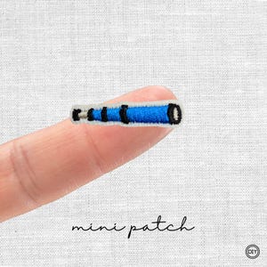 Könnte beinhalten: Ein kleiner gestickter Aufnäher in Form eines Fernrohrs mit einem leuchtend blauen Körper, schwarzen Akzenten und einem weißen Rand. Das Design ist mit horizontalen Stichen und einer klaren, zylindrischen Form versehen, präsentiert als Miniatur-Accessoire.