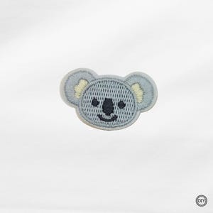 Könnte beinhalten: Ein hellblau gestickter Koala-Aufnäher mit einem lächelnden Gesicht, schwarzen Augen und gelben Akzenten an den Ohren. Ideal für DIY-Projekte und Bastelarbeiten.
