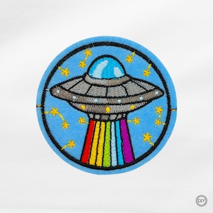 Könnte beinhalten: Gestickter Aufnäher mit einer grauen und silbernen UFO mit einer blauen Kuppel, die einen Regenbogenstrahl aussendet. Das UFO befindet sich vor einem hellblauen Hintergrund mit gelben Sternakzenten, alles innerhalb eines schwarzen kreisförmigen Randes. Ein DIY-Aufnäher.