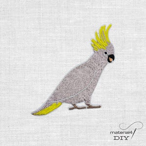 Könnte beinhalten: Gestickter Aufnäher mit einem grauen Kakadu mit gelbem Kamm und Schwanzfedern. Der Vogel hat einen schwarzen Schnabel und sitzt auf weißem Hintergrund. Geeignet für DIY-Projekte oder Kleidung.