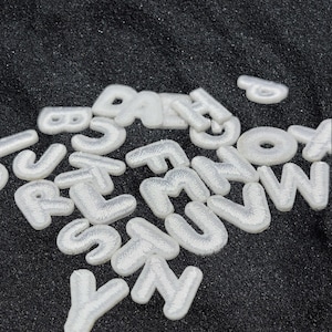 Könnte beinhalten: Eine Sammlung weißer, gestickter Buchstaben-Aufnäher mit einer strukturierten, erhabenen Stickerei. Jeder einzelne Buchstabe ist mit sauberem weißem Garn gefertigt und bildet einen deutlichen Kontrast zur dunklen, körnigen Oberfläche.