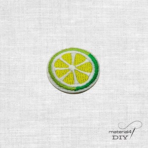 Könnte beinhalten: Ein kreisförmiger Stickaufnäher, der einen Querschnitt einer Limette darstellt. Das Design zeigt ein hellgelbes Zentrum mit strahlenförmigen Segmenten, umgeben von einem dicken, strukturierten grünen Rand, der die Schale imitiert.