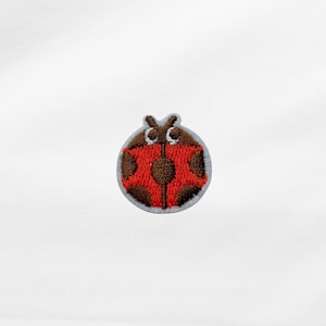Könnte beinhalten: Gestickter Marienkäfer-Aufnäher mit rotem Körper, braunen Flecken und einem braunen Kopf. Der Aufnäher hat einen weißen Rand und ist zum Aufnähen oder Aufbügeln auf Stoff vorgesehen. Geeignet für Kleidung oder Accessoires.