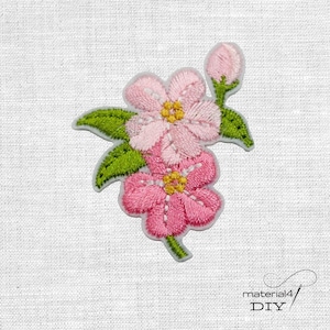 Könnte beinhalten: Gestickter Blumenaufnäher mit zwei rosa Blüten mit gelben Zentren und grünen Blättern. Eine Knospe ist ebenfalls sichtbar. Der Aufnäher befindet sich auf weißem Hintergrund. Der Text "material4 DIY" befindet sich unten rechts.