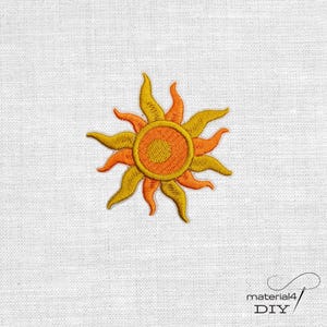 Könnte beinhalten: Gesticktes Sonnen-Patch in Gelb-, Orange- und Goldtönen. Die Sonne hat eine kreisförmige Mitte und spitze Strahlen. Das Patch befindet sich auf einem weißen Stoffhintergrund. Der Text "material4 DIY" befindet sich in der unteren rechten Ecke.