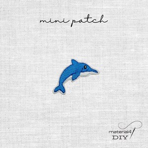 Könnte beinhalten: Ein blaues, gesticktes Delfin-Patch mit weißem Rand. Das Patch hat ein kleines Auge und befindet sich auf einem weißen Hintergrund. Über dem Delfin steht in Schreibschrift "mini patch". Darunter steht "material4 DIY".