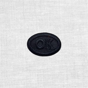 Könnte beinhalten: Ein marineblauer, gestickter Aufnäher in ovaler Form. Der Aufnäher zeigt die Buchstaben "OK" in einer stilisierten Schriftart. Der Aufnäher ist vor einem weißen, strukturierten Hintergrund platziert, wodurch ein Kontrast zwischen dem dunklen Aufnäher und dem hellen Hintergrund entsteht.